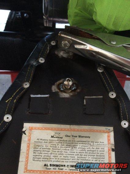 Underside of a black motorcycle seat with yellow stitching, showing a metal bracket and a warranty tag