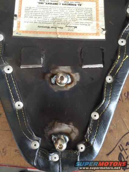 Underside of a black motorcycle seat with yellow stitching and metal fasteners. A warranty tag is visible