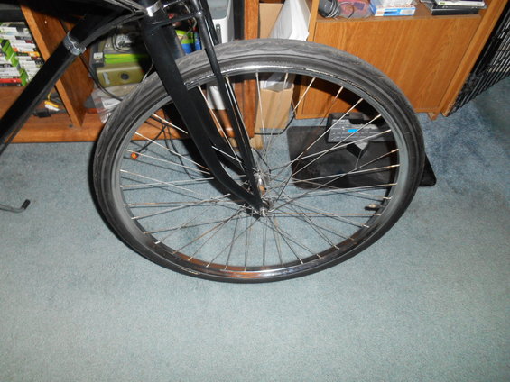 Close-up of a bicycle's front wheel and fork, with spokes and tire visible. A wooden shelf unit is in the background