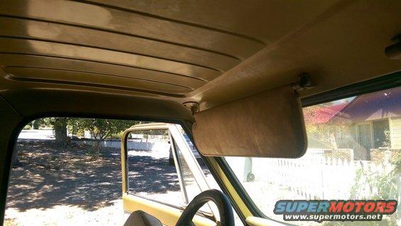 Yellow truck interior showing a tan headliner, sun visor, and steering wheel. Exterior trees and houses visible through the open door and window