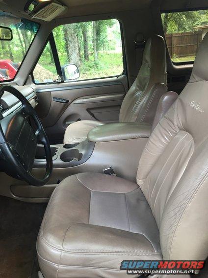 Interior view of a Ford Bronco, showing tan leather seats, center console with cup holders, and steering wheel. Trees visible through the windows