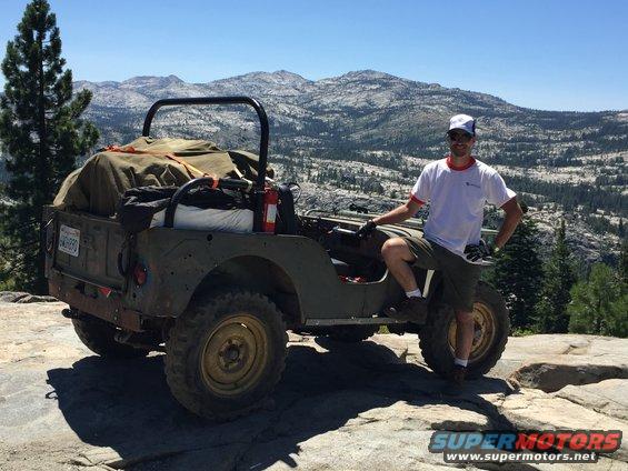 Man posing with a green Willys CJ-3A loaded with gear on a rocky mountain overlook