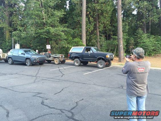 Man photographs a dark Ford Bronco and a vintage Jeep trailer parked in a lot