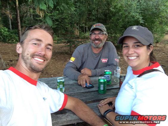 Three smiling people are sitting at a picnic table outdoors with drinks