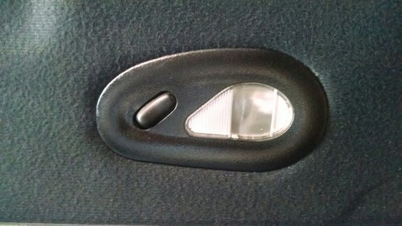 Black oval interior dome light with a push button and a clear ribbed lens, mounted on dark grey fabric