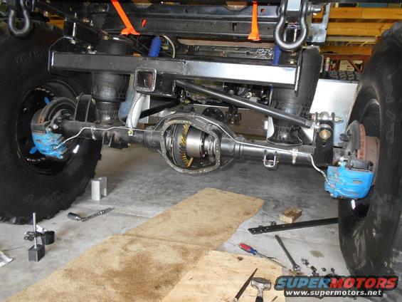 Rear axle assembly with blue brake calipers and large tires, undergoing maintenance