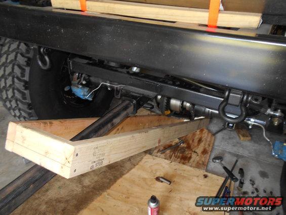 Close-up of vehicle undercarriage, showing frame, suspension, and a tire. Wooden blocks support the vehicle