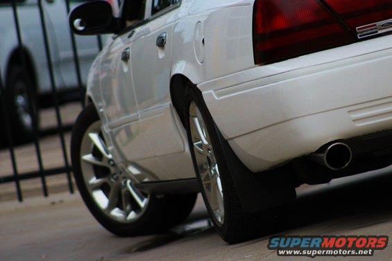 White sedan's rear quarter panel and wheel. Another car is visible behind a fence