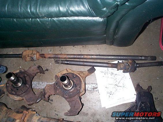 Two front axle knuckles and driveshafts laid out on the floor next to a green couch