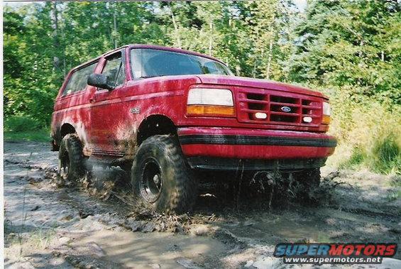 1995 Ford Bronco driving through a muddy trail with trees in the background