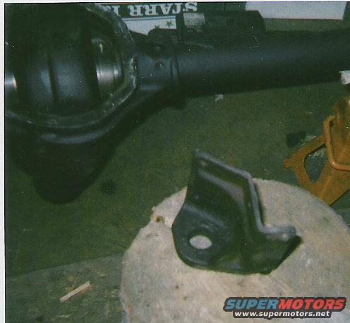 Vehicle rear axle assembly and a metal bracket on a block. A yellow jack stand is in the background