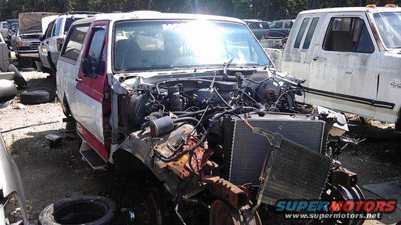 Junkyard Bronco with front end removed, showing engine bay and radiator. Other vehicles in background