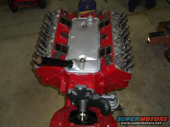 V8 engine with red block, silver intake manifold, and exposed valve springs. Water pump and carburetor visible