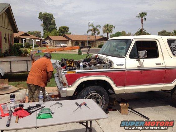Man working on the engine of a red and white 1978 Ford Bronco, with tools laid out on a table