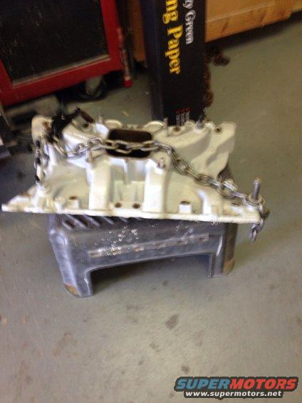 White engine intake manifold with a metal chain draped over it, resting on a gray stand