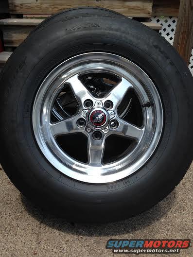 Chrome five-spoke wheel with a black tire, center cap has red and black logo