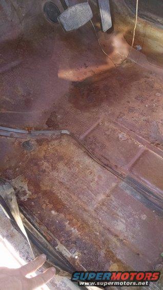 Rusted floor pan with brake and accelerator pedals visible. A hand is touching the lower left edge
