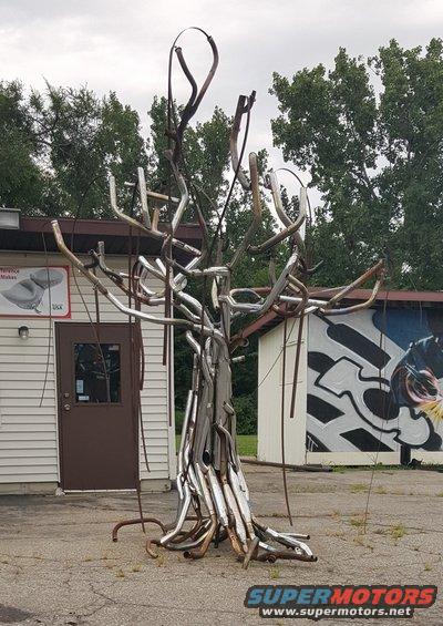 Exhaust pipe sculpture resembling a tree in front of a building with a mural