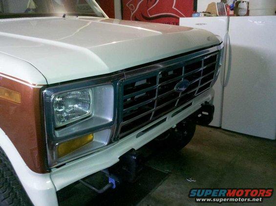 Close-up of a white Ford Bronco's front grille and headlight. The grille has horizontal bars and a blue Ford emblem