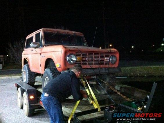 Orange 1974 Ford Bronco on a trailer at night, being secured with straps