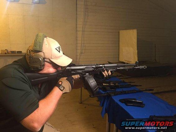 Man wearing a cap and ear protection aims a rifle with a drum magazine, with other firearms on a table behind him