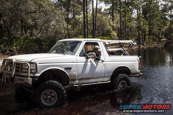White Ford Bronco with a soft top driving through shallow water, front bull bar visible