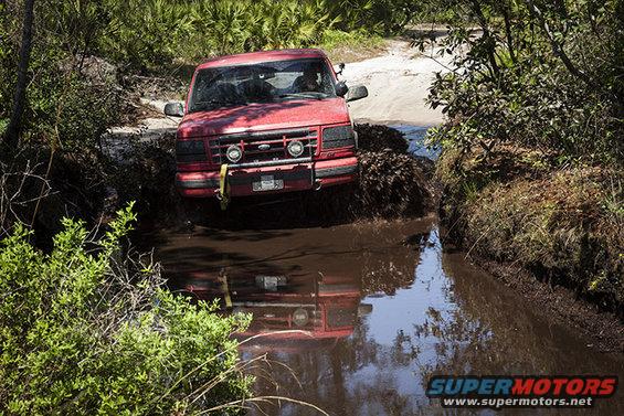 Red 1995 Ford Bronco driving through a muddy ditch with trees on either side
