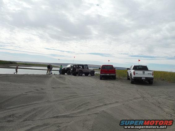 2005 Jeep Wrangler parked on sand near water, with red and white pickup trucks behind it