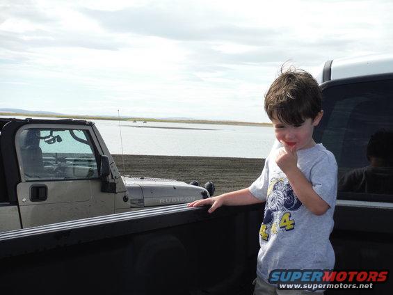 Boy in a pickup truck bed looking at a Jeep Wrangler parked by a body of water