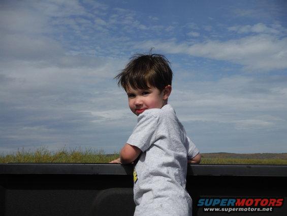 Child in gray t-shirt leans over dark truck bed railing, looking back at camera. Blue sky with clouds above