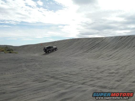 Gray Jeep Wrangler driving up a sand dune under a cloudy sky