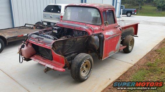 Red pickup truck missing hood and fenders, sitting on a concrete pad next to a trailer