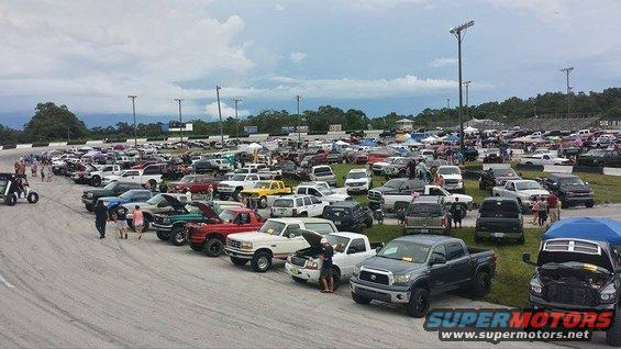 Lots of trucks and SUVs parked in a lot, some with hoods up. People milling around