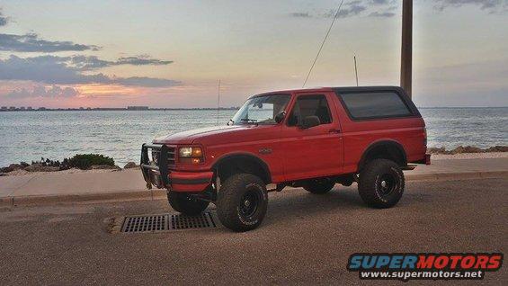 Red Ford Bronco parked by the water at sunset. It has a bull bar on the front