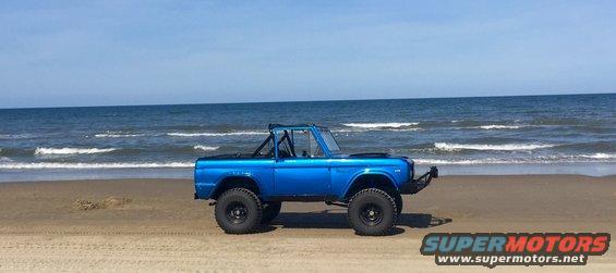 Blue 1974 Ford Bronco with a roll cage parked on a sandy beach with the ocean in the background