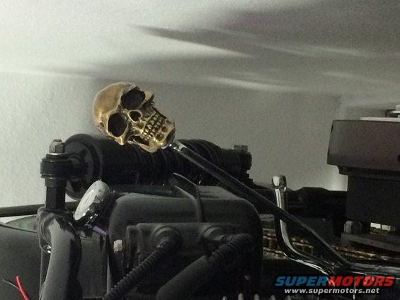 Close-up of a motorcycle engine component with a brass skull ornament on top. A gauge is visible on the left