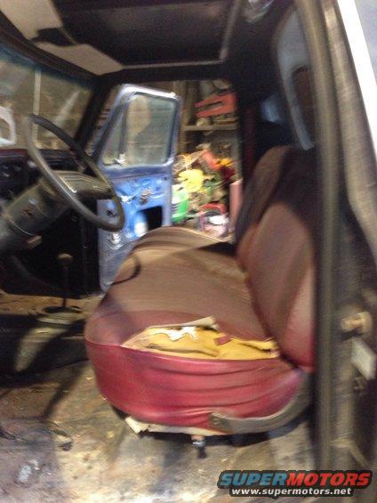 Interior of a vehicle with torn red and tan seats, a black steering wheel, and an open blue door. Tools are visible in the background