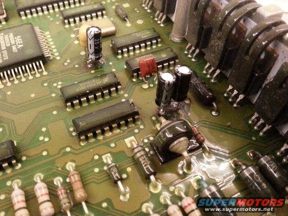 Close-up of a green circuit board with various electronic components including integrated circuits, capacitors, and resistors. Some text is visible on the chips