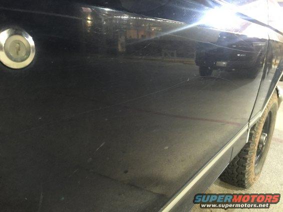 Black vehicle side panel with a lock cylinder and visible scratches. Tire and part of the rear fender are visible