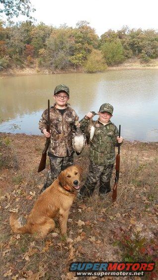 Two boys in camouflage with hunting rifles and a dog pose by a lake with their duck catch