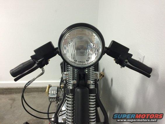 Motorcycle front end with black handlebars, large round headlight, and exposed springs. A small skull charm hangs on the left