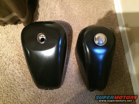Two black motorcycle gas tanks sit on a carpeted floor. One has a chrome gas cap, the other has an open fuel filler neck