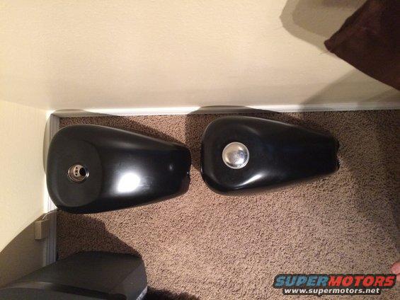 Two black motorcycle fuel tanks, one with a silver cap, resting on a carpeted floor