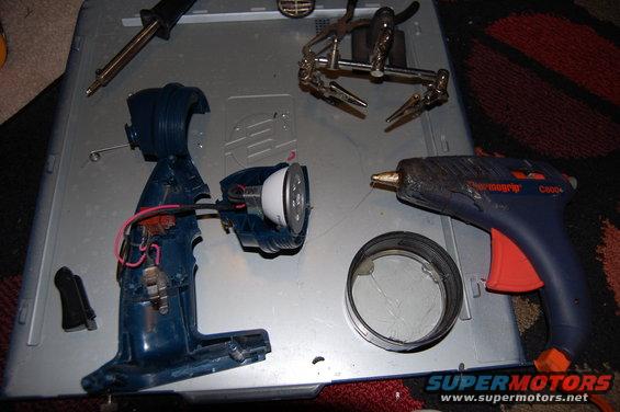 Disassembled blue flashlight with wires exposed, next to a hot glue gun, soldering iron, and helping hands tool