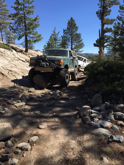 Green off-road SUV with large tires navigating a rocky trail, another white SUV behind it
