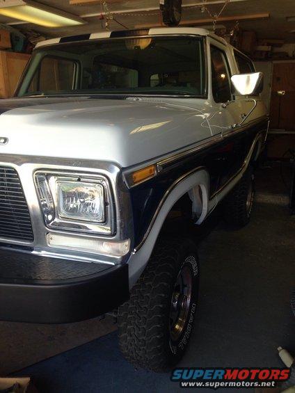 Front 3/4 view of a 1978 Ford Bronco in a garage. It has a two-tone paint job with a dark blue lower half and a silver upper half, with a black and white striped roof