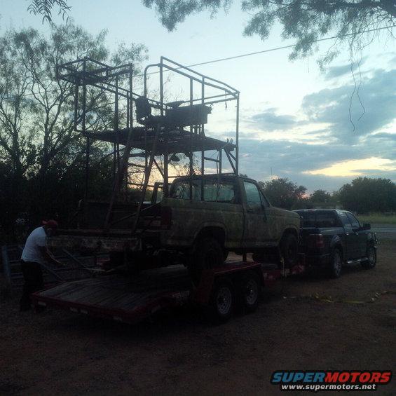 Green Ford F-150 on a trailer, loaded with a tall metal frame, parked next to a dark pickup truck