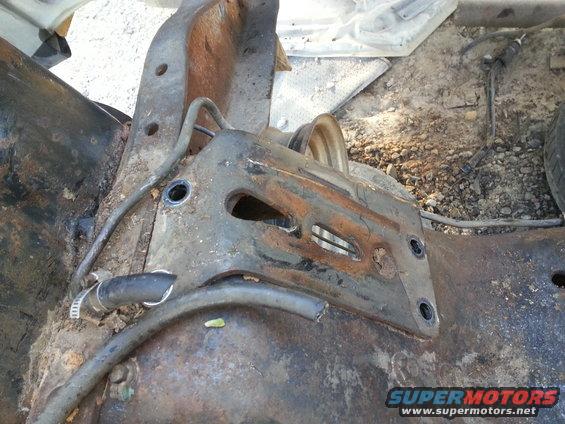 Rusted vehicle frame section with hoses and a pulley wheel, showing significant corrosion