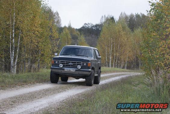 Dark blue 1987 Ford Bronco driving on a dirt road with autumn trees