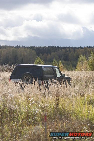 1987 Ford Bronco parked in tall dry grass with trees and cloudy sky in background
